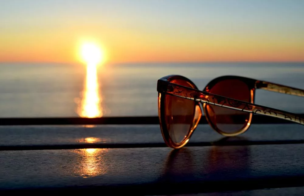 A pair of sunglasses sitting on table at the ocean shore during sunset.