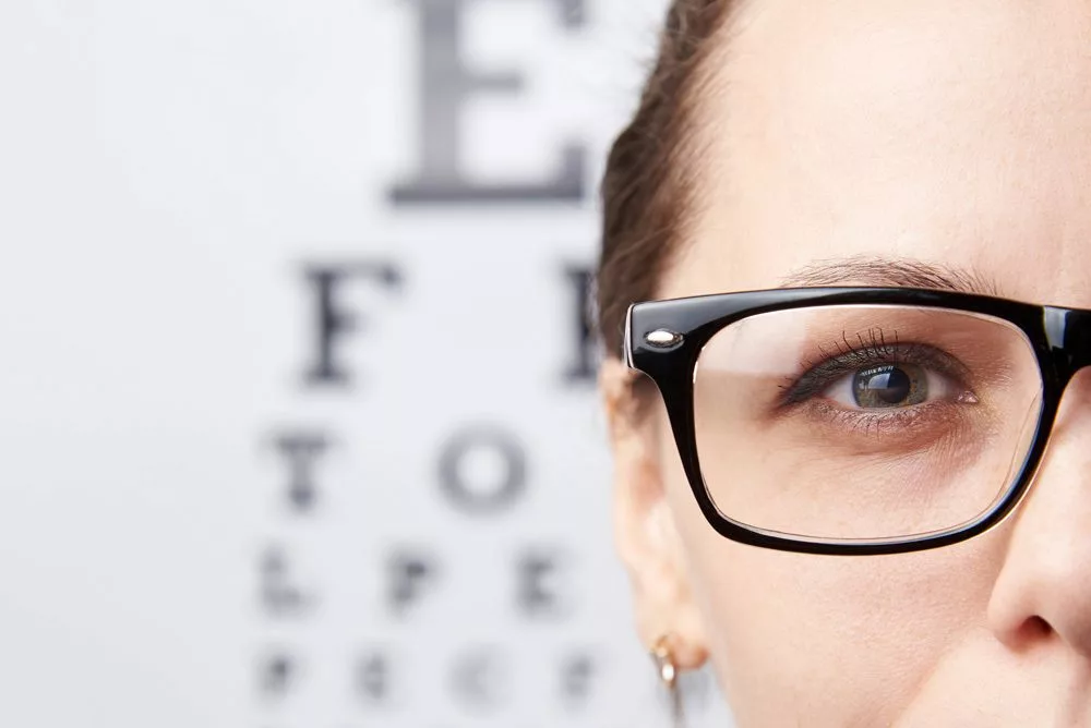 Woman with glasses standing in front of Snellen eye chart.