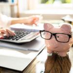 person Calculating Bill In Front Of Pink Piggy Bank with glasses on Over Desk