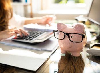 person Calculating Bill In Front Of Pink Piggy Bank with glasses on Over Desk