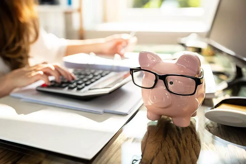 person Calculating Bill In Front Of Pink Piggy Bank with glasses on Over Desk