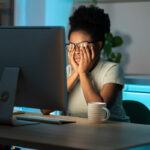 African American woman rubbing her eyes under her glasses while sitting in the dark at a computer