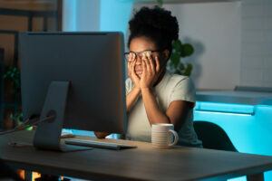 African American woman rubbing her eyes under her glasses while sitting in the dark at a computer