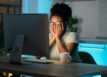 African American woman rubbing her eyes under her glasses while sitting in the dark at a computer