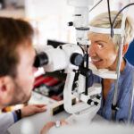 Gray-haired, smiling woman with on chin rest in eye exam with young male doctor