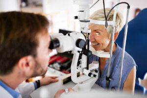 Gray-haired, smiling woman with on chin rest in eye exam with young male doctor