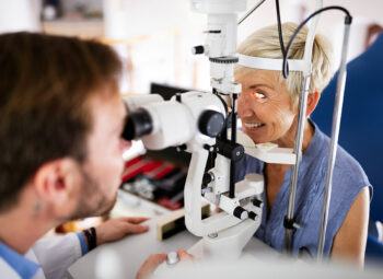 Gray-haired, smiling woman with on chin rest in eye exam with young male doctor