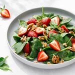 strawberry spinach and walnut salad in white bowl with ingredients also scattered nearby