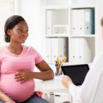 Smiling pregnant African American woman in pink top holding her hand on her belly during consult with female doctor