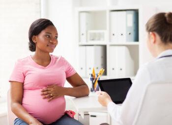 Smiling pregnant African American woman in pink top holding her hand on her belly during consult with female doctor