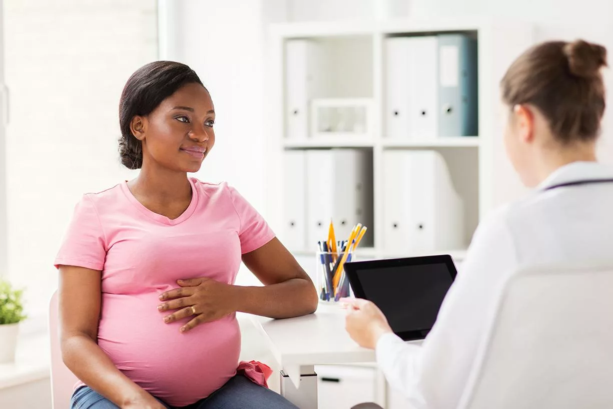 Smiling pregnant African American woman in pink top holding her hand on her belly during consult with female doctor