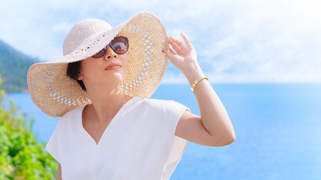 woman in sunglasses on a sunny day near a lake or ocean wearing and holding the brim of a wide-brimmed hat