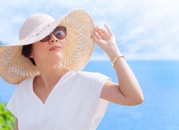 woman in sunglasses on a sunny day near a lake or ocean wearing and holding the brim of a wide-brimmed hat