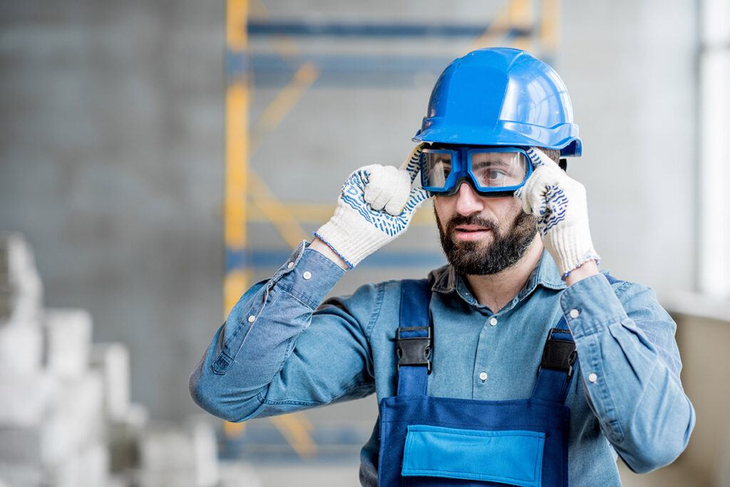 bearded workman holding gloved hands to the safety glasses he wears under his hard hat