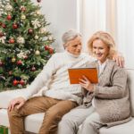 senior couple on couch by Christmas tree, looking at a tablet