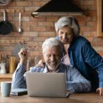 Grey-haired, smiling man and woman looking at laptop, with man holding glasses rather than wearing them.