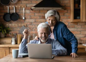 Grey-haired, smiling man and woman looking at laptop, with man holding glasses rather than wearing them.