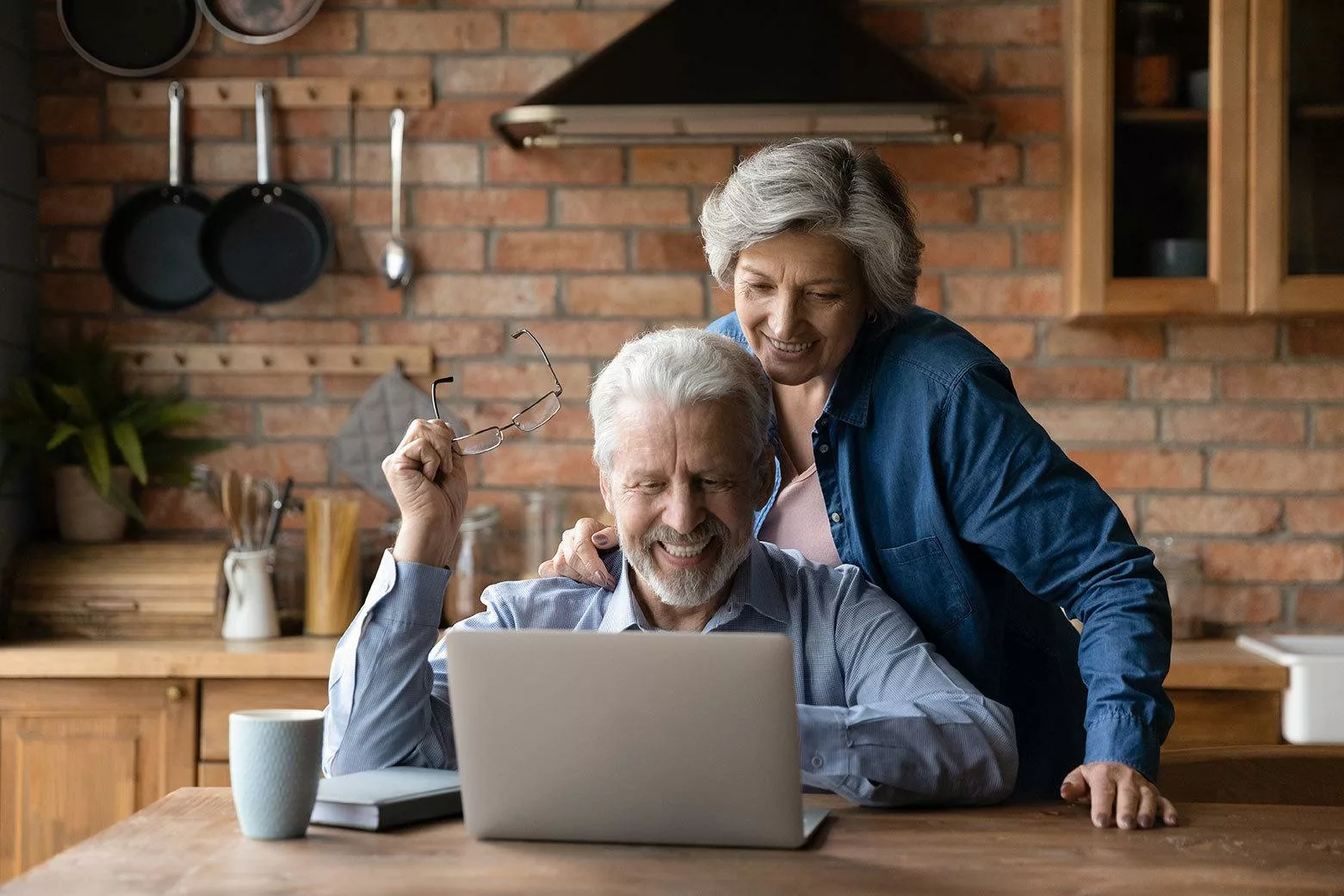 Grey-haired, smiling man and woman looking at laptop, with man holding glasses rather than wearing them.