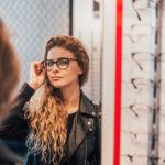 Long-haired woman in leather jacket trying on glass frames while looking in mirror