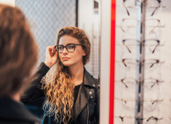 Long-haired woman in leather jacket trying on glass frames while looking in mirror