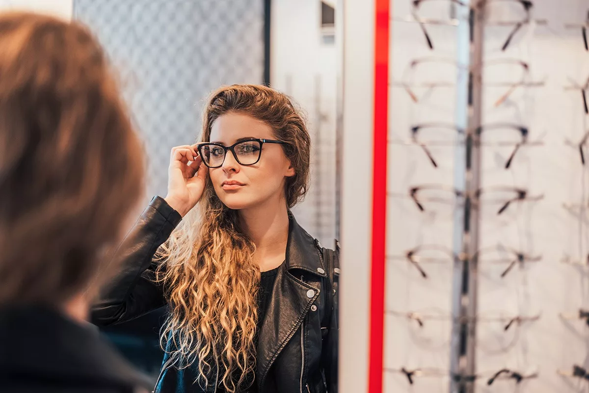 Long-haired woman in leather jacket trying on glass frames while looking in mirror