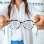 Torso of woman in coat standing in front of eyeglasses display, holding out a pair of glasses earpieces first