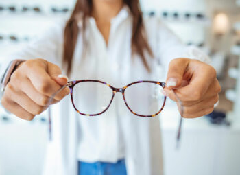 Torso of woman in coat standing in front of eyeglasses display, holding out a pair of glasses earpieces first