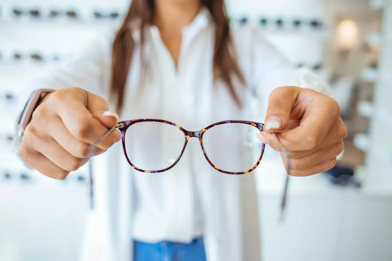 Torso of woman in coat standing in front of eyeglasses display, holding out a pair of glasses earpieces first