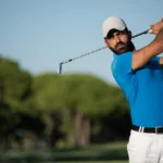 pro golfer in white ball cap and pants, and blue polo shirt, immediately after swing, watching ball.