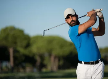 pro golfer in white ball cap and pants, and blue polo shirt, immediately after swing, watching ball.