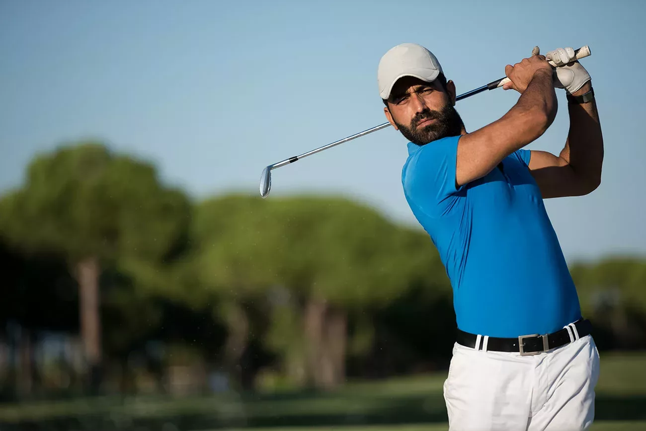 pro golfer in white ball cap and pants, and blue polo shirt, immediately after swing, watching ball.