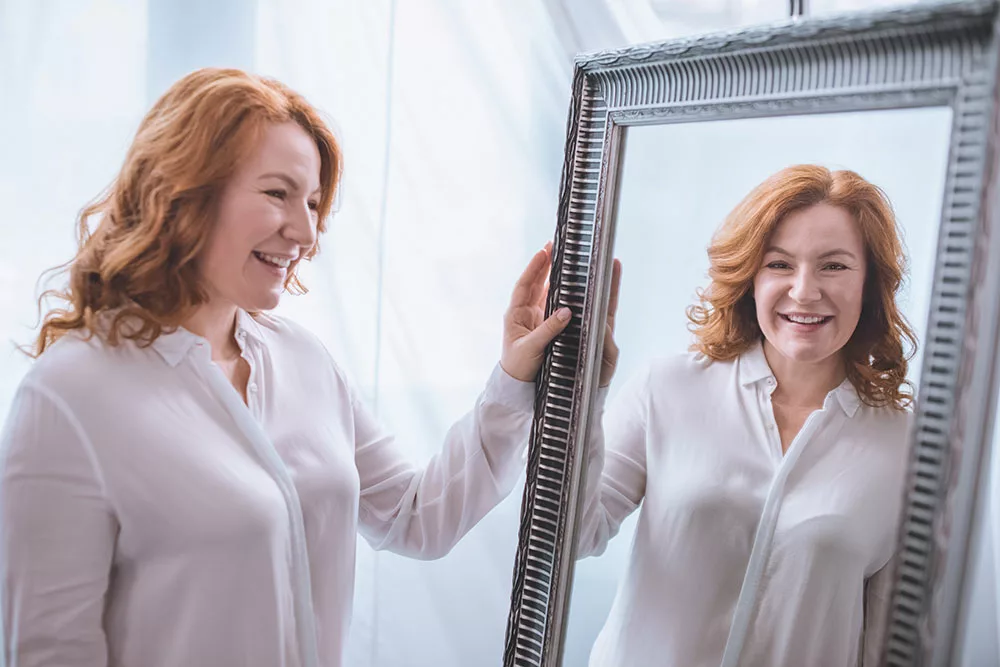 Redheaded woman in her 30s smiling at her reflection in a mirror