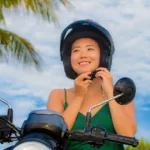 Young woman on scooter against partly cloudy blue sky and palm tree adjusting strap on helmet.