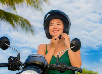 Young woman on scooter against partly cloudy blue sky and palm tree adjusting strap on helmet.