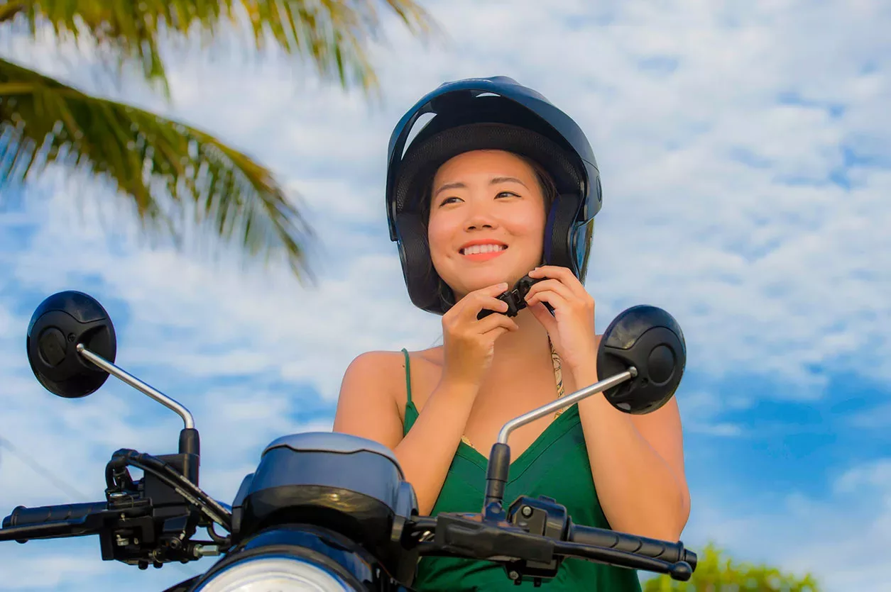 Young woman on scooter against partly cloudy blue sky and palm tree adjusting strap on helmet.