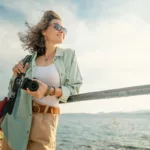 Woman in sunglasses holding camera at railing near body of water on a sunny day.