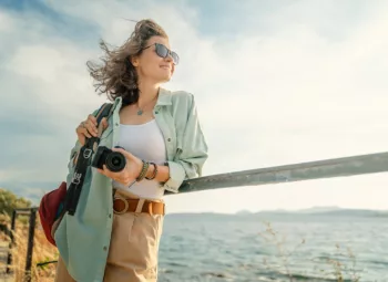 Woman in sunglasses holding camera at railing near body of water on a sunny day.