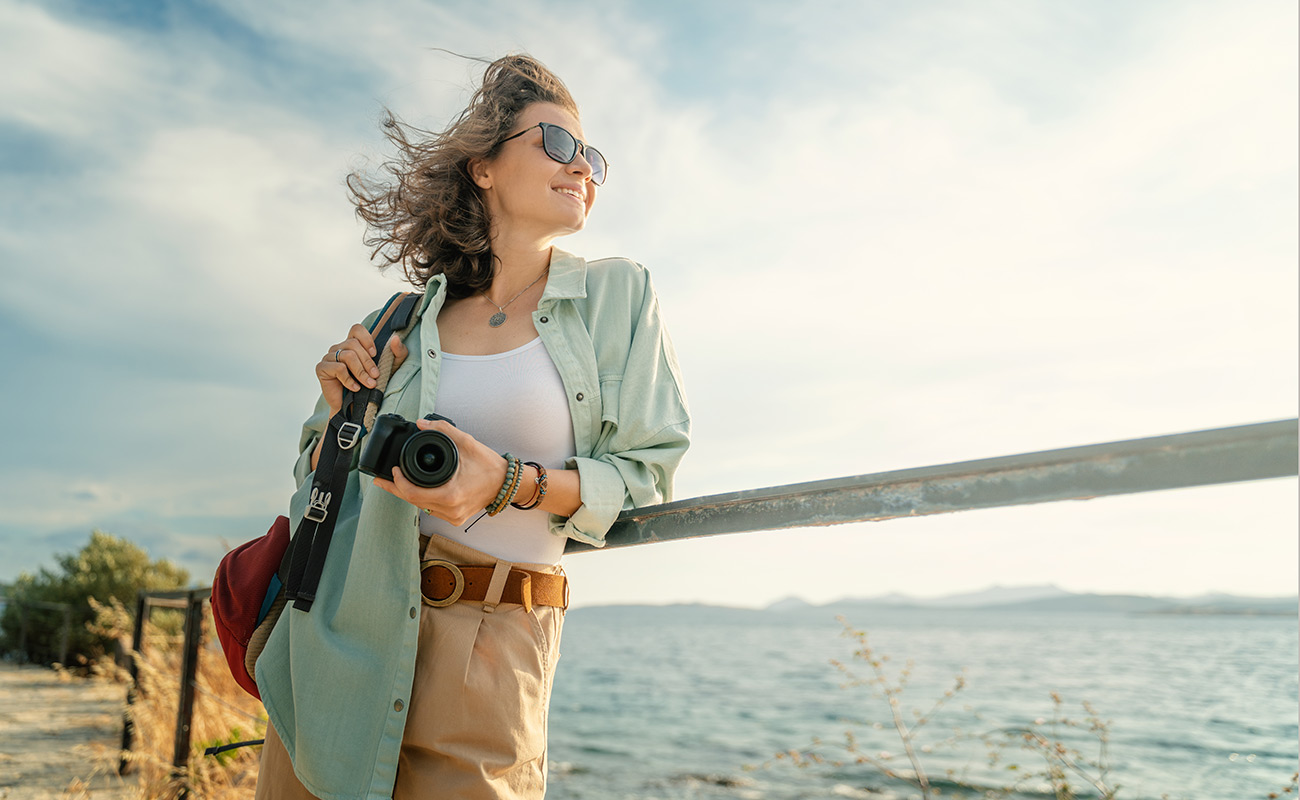 Woman in sunglasses holding camera at railing near body of water on a sunny day.