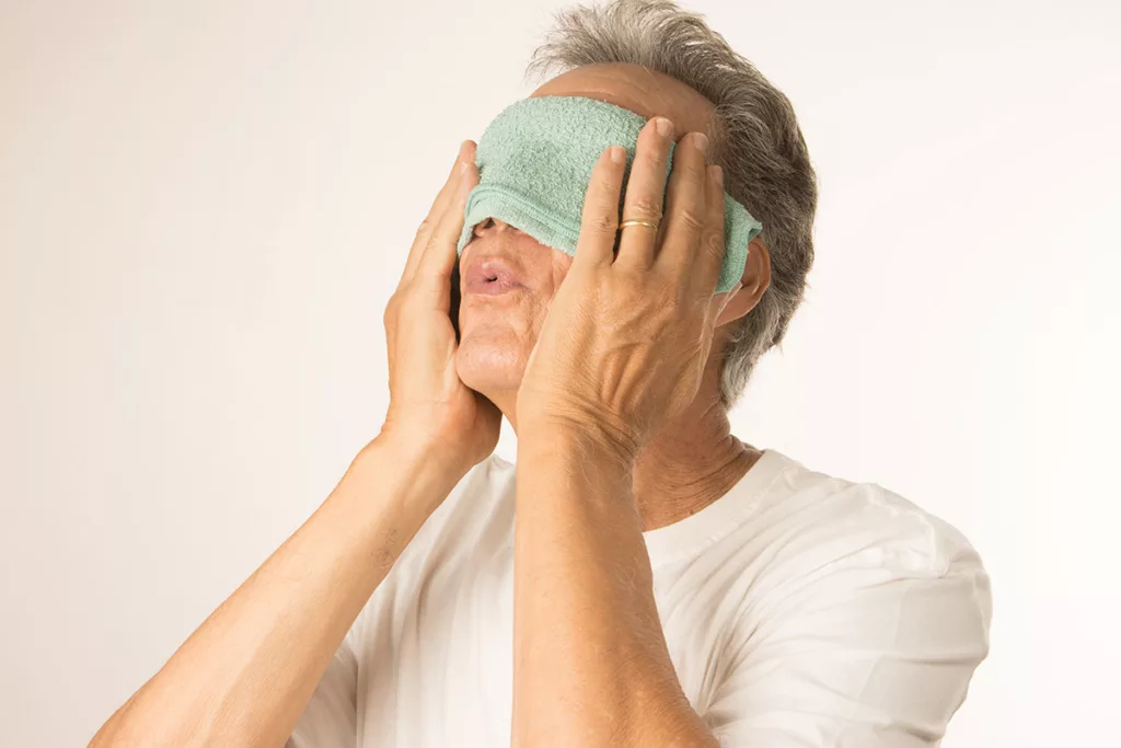 man holding washcloth over his eyes and while making an ooh face with his lips