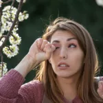woman rubbing eye in front of blossoming tree