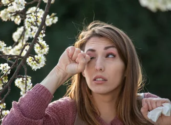 woman rubbing eye in front of blossoming tree