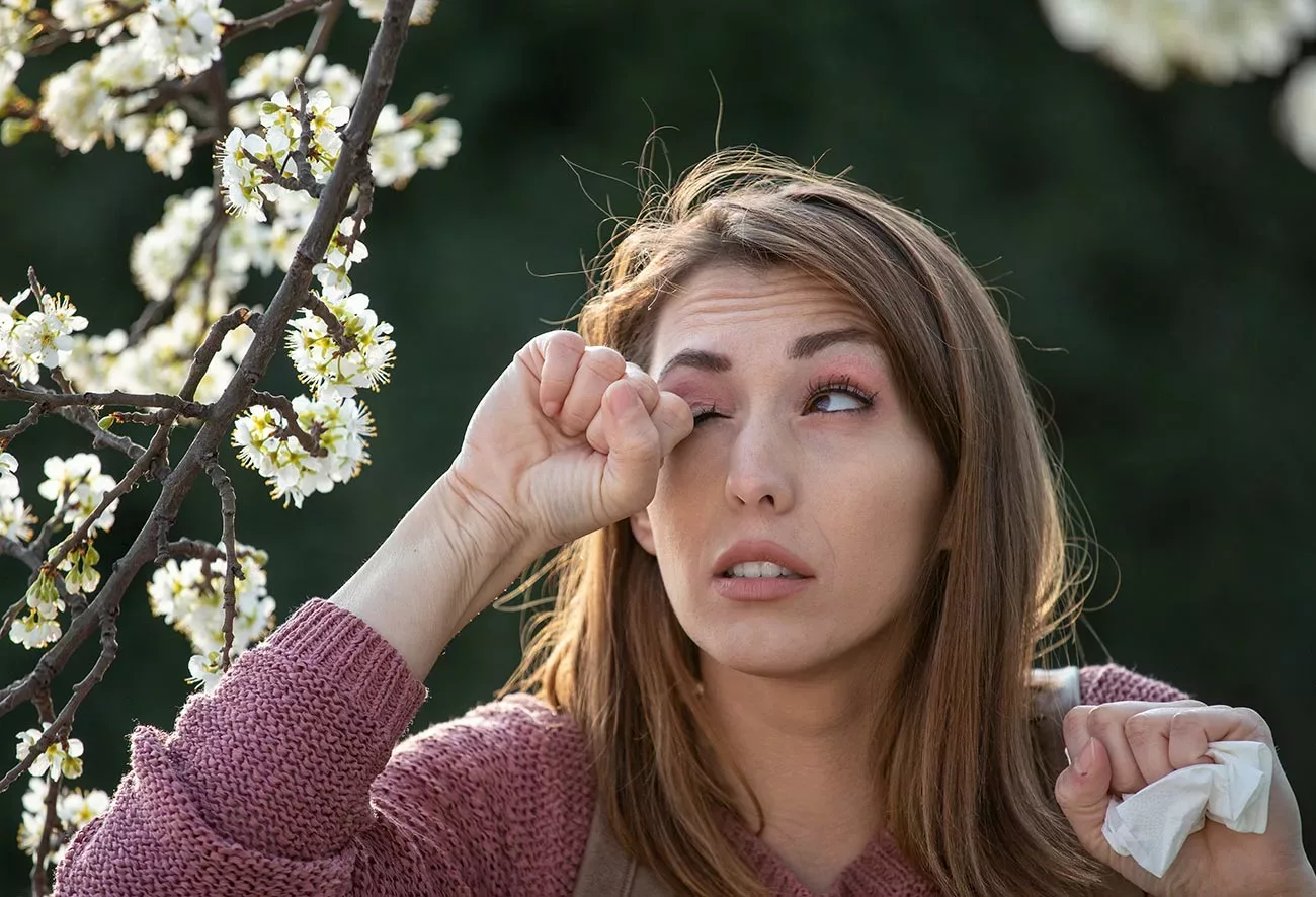 woman rubbing eye in front of blossoming tree