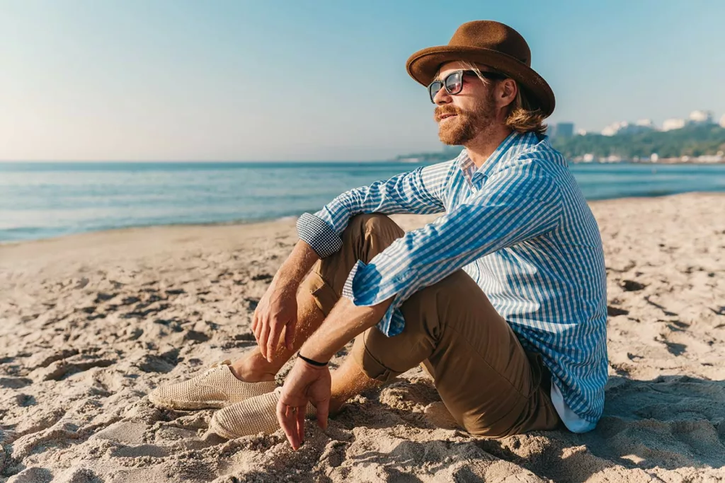 Bearded redheaded man wearing hat and sunglasses sitting on a beach. 