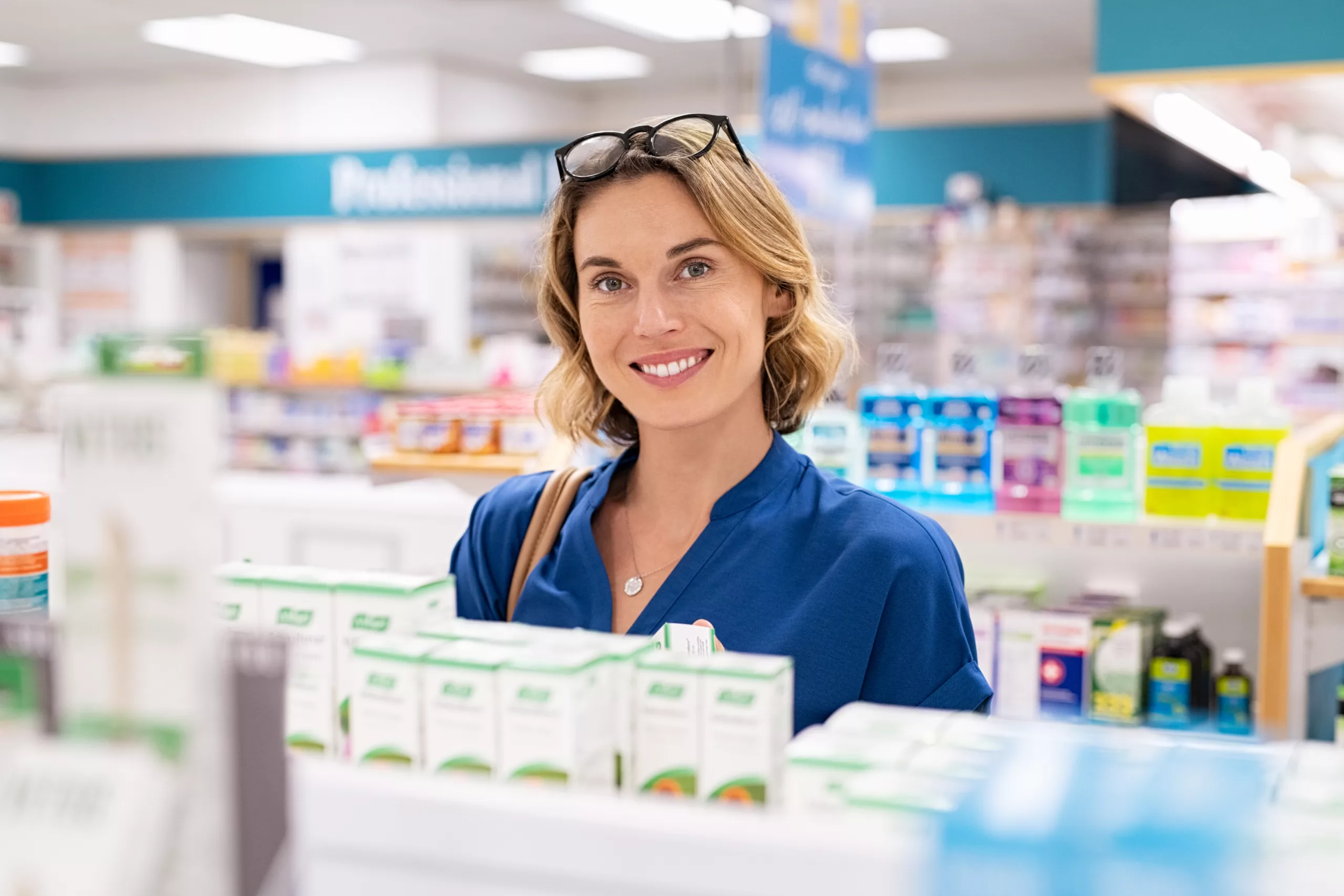 Portrait of smiling woman choosing dietary supplement at pharmacy in shopping mall. Happy mature woman customer buying lotion in skincare section of chemist’s. Woman checking medicine and drugs in shelf at drugstore while looking at camera.