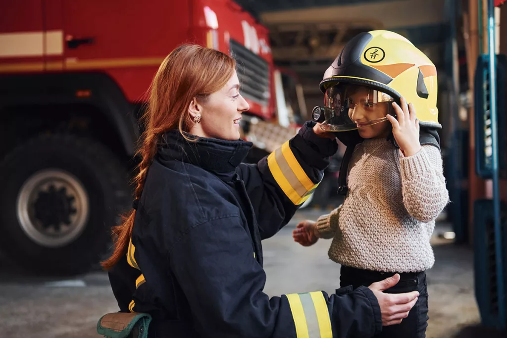 A female firefighter first responder interacts with a young girl in a helmet.