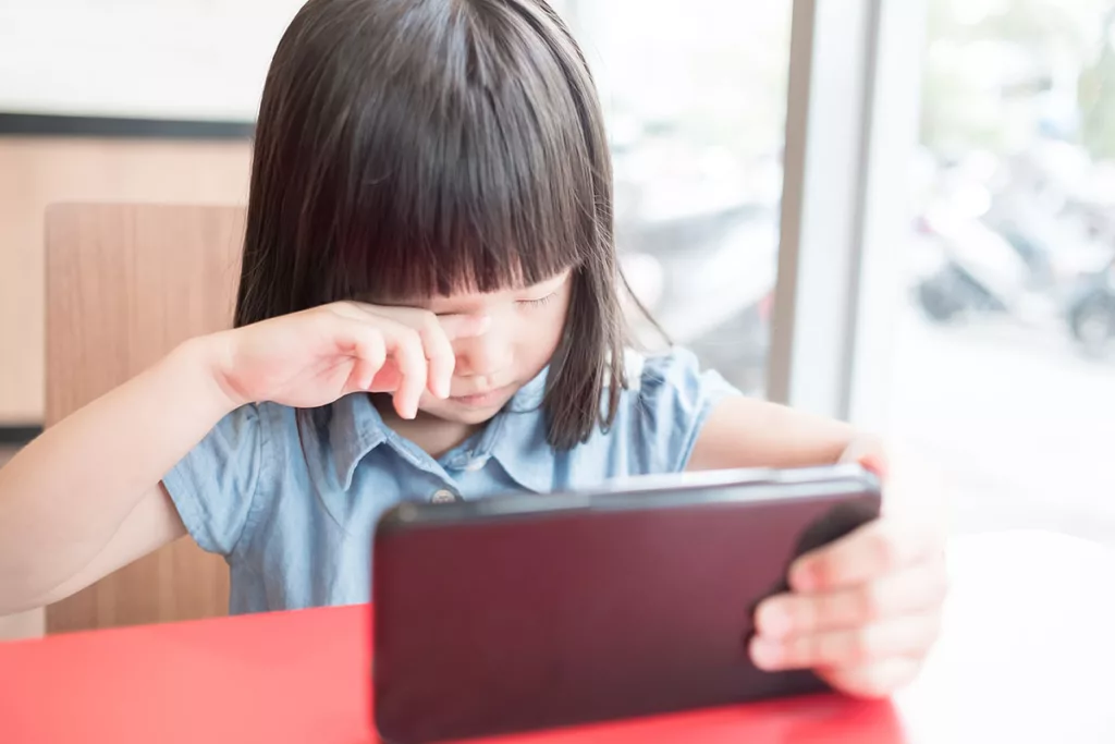 A young girl experiencing light sensitivity rubs her eyes while looking at a tablet.