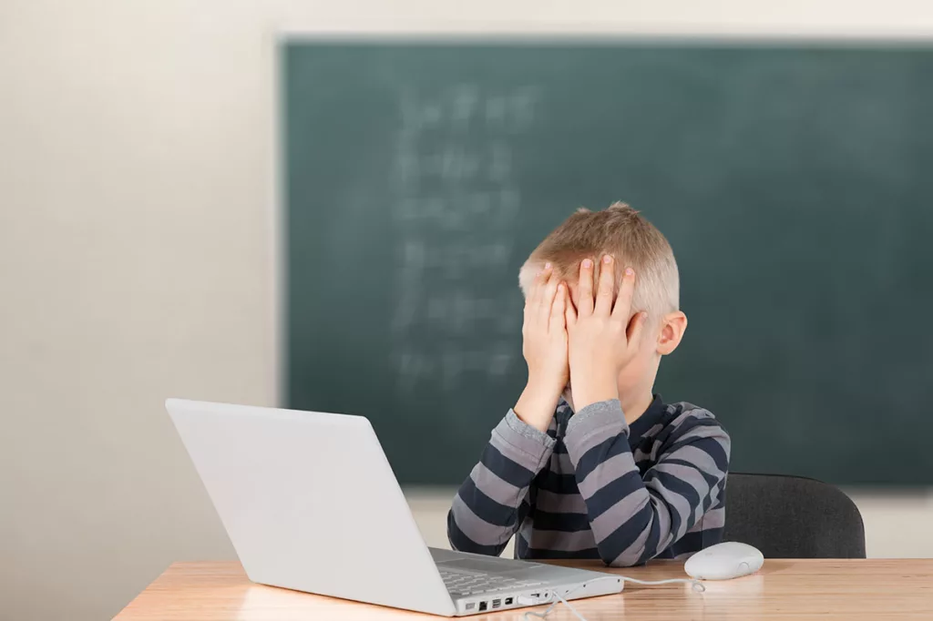 A young child sitting at a desk in front of a chalkboard is struggling in school due to vision concerns.