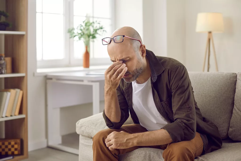 A man sits on a couch and rubs his eyes due to an ocular migraine.