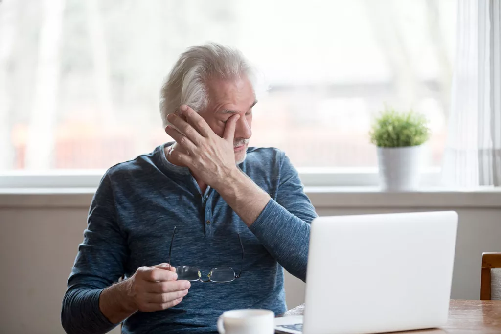 A man sitting by a laptop experiencing dry eye rubs his eye.