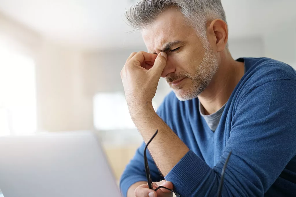 A man with cataracts sits in front of a laptop, rubbing his eyes due to light sensitivity.